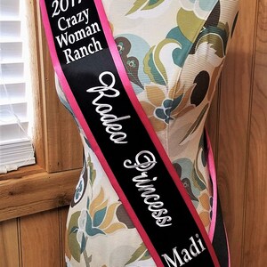May include: A black and pink sash with the text "2017 Crazy Woman Ranch Rodeo Princess Madi" in white lettering.
