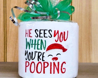 May include: A white roll of toilet paper wrapped in green and silver cellophane with a red and white Santa hat and the text "He sees you when you're pooping" embroidered on the front.
