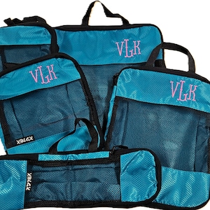 May include: A set of four teal packing cubes with black trim and mesh panels. Each bag features a pink embroidered monogram reading VLK and a small black tag with the white text XBLGX. The bags have black carrying handles and zipper closures.