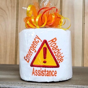 May include: A white toilet paper roll with a red and yellow embroidered warning sign that says "Roadside Assistance" and "Emergency". The toilet paper roll is wrapped in orange and yellow ribbon.