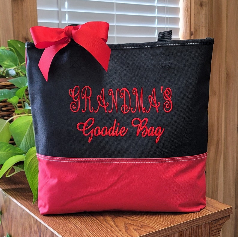 Puede incluir: Bolsa de lona negra con un lazo rojo y texto bordado en rojo que dice "Grandma's Goodie Bag". La bolsa tiene un fondo rojo.