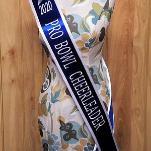 K&ouml;nnte beinhalten: Ein wei&szlig;es und blaues Satinsband mit dem Text "2020 PRO BOWL CHEERLEADER" in silbernen Buchstaben. Ein blauer Stern befindet sich auf der linken Seite des Bandes.