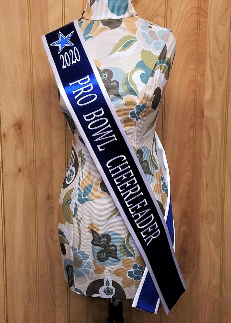 K&ouml;nnte beinhalten: Ein wei&szlig;es und blaues Satinsband mit dem Text "2020 PRO BOWL CHEERLEADER" in silbernen Buchstaben. Ein blauer Stern befindet sich auf der linken Seite des Bandes.