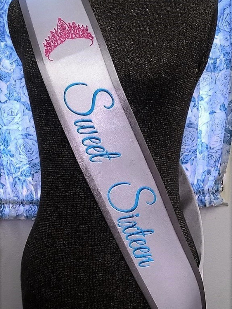 May include: A white satin sash with a silver trim and blue embroidery that reads "Sweet Sixteen". A pink crown is embroidered at the top of the sash.