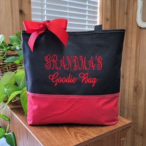Puede incluir: Bolsa de tela negra y roja con un lazo rojo en la parte superior. La bolsa tiene el texto "Grandma's Goodie Bag" bordado en rojo en la parte delantera.
