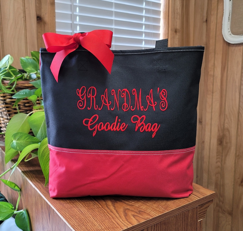 Puede incluir: Bolsa de tela negra y roja con un lazo rojo en la parte superior. La bolsa tiene el texto "Grandma's Goodie Bag" bordado en rojo en la parte delantera.