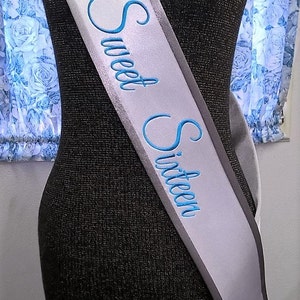 May include: A white satin sash with a gray border and blue embroidery that reads "Sweet Sixteen". A pink crown is embroidered on the sash.