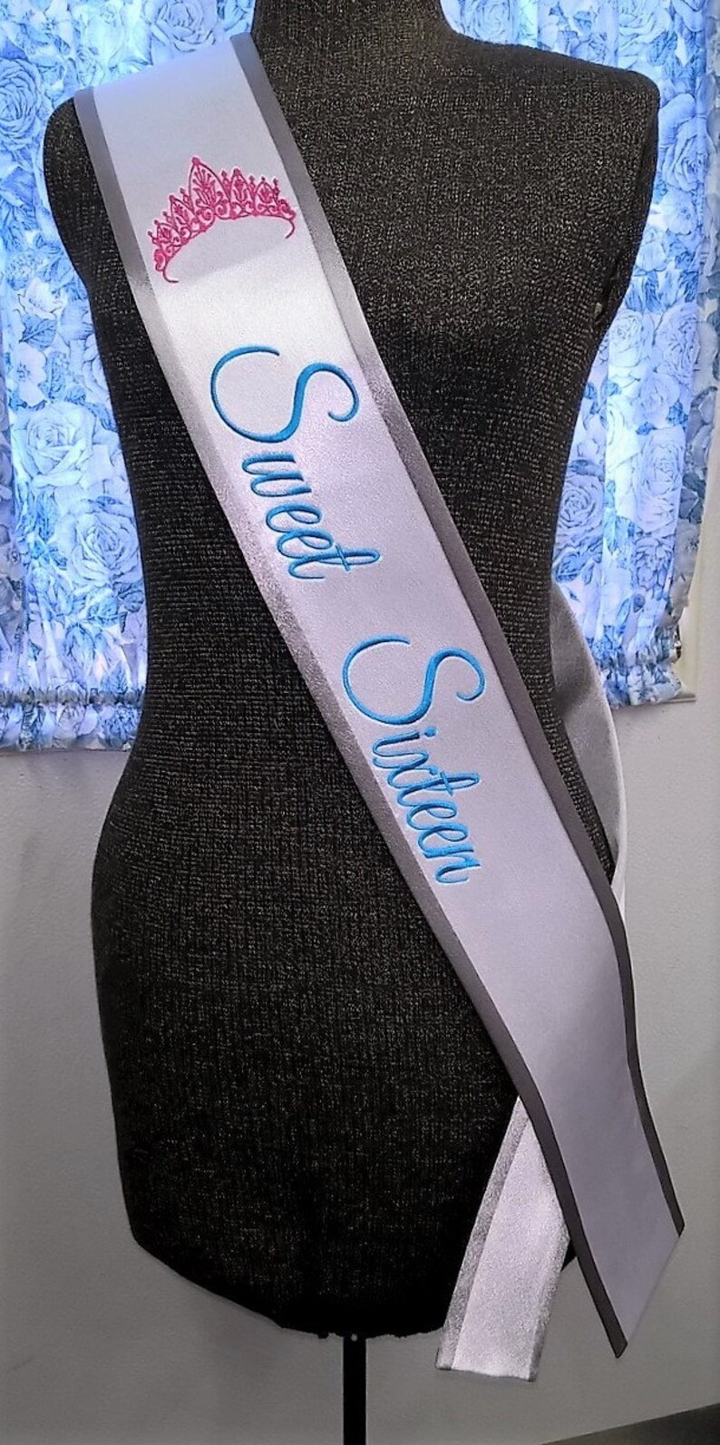 May include: A white satin sash with a gray border and blue embroidery that reads "Sweet Sixteen". A pink crown is embroidered on the sash.