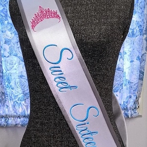 May include: A white satin sash with a silver border and blue embroidery that reads "Sweet Sixteen". A pink crown is embroidered at the top of the sash.