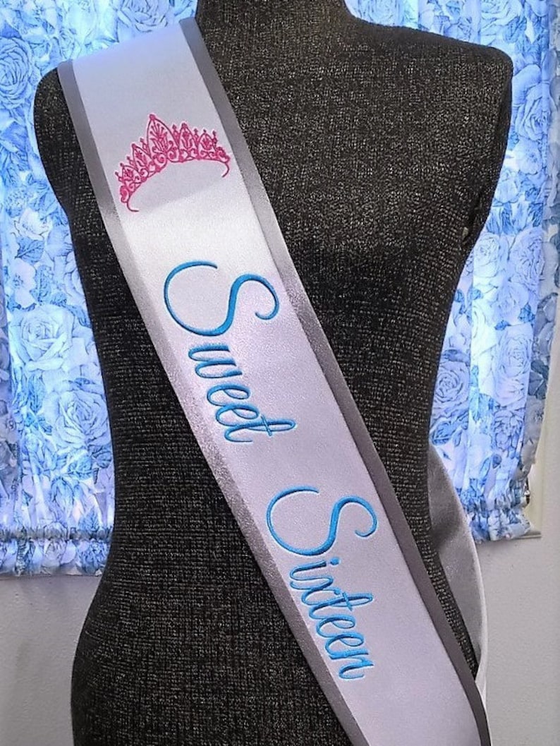 May include: A white satin sash with a silver border and blue embroidery that reads "Sweet Sixteen". A pink crown is embroidered at the top of the sash.