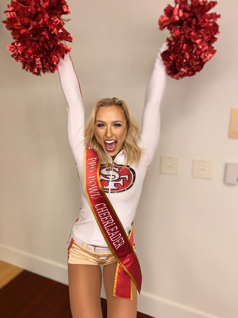 K&ouml;nnte beinhalten: Eine Frau in einem wei&szlig;en Cheerleader-Outfit mit einem roten Band, auf dem "Pro Bowl Cheerleader" und ein goldenes und rotes Logo stehen. Sie h&auml;lt rote Pompons in jeder Hand und l&auml;chelt.