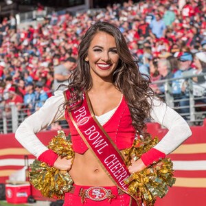 K&ouml;nnte beinhalten: Eine Frau in einem rot-wei&szlig;en Cheerleader-Outfit mit einer goldenen Sch&auml;rpe, auf der "Pro Bowl Cheerleader" steht. Sie h&auml;lt zwei goldene Pompons und l&auml;chelt in die Kamera.