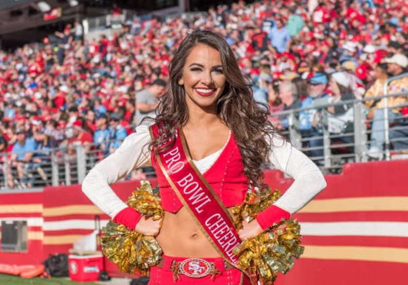 K&ouml;nnte beinhalten: Eine Frau in einem rot-wei&szlig;en Cheerleader-Outfit mit einer goldenen Sch&auml;rpe, auf der "Pro Bowl Cheerleader" steht. Sie h&auml;lt zwei goldene Pompons und l&auml;chelt in die Kamera.