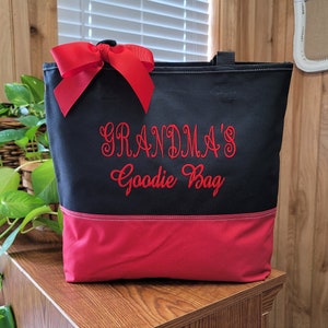 Puede incluir: Bolsa de lona negra con un lazo rojo y un dise&ntilde;o bordado en rojo que dice "Grandma's Goodie Bag". La bolsa tiene un fondo rojo.