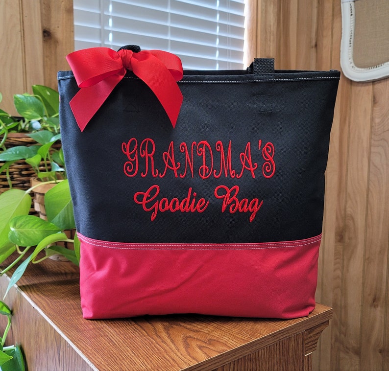 Puede incluir: Bolsa de lona negra con un lazo rojo y un dise&ntilde;o bordado en rojo que dice "Grandma's Goodie Bag". La bolsa tiene un fondo rojo.