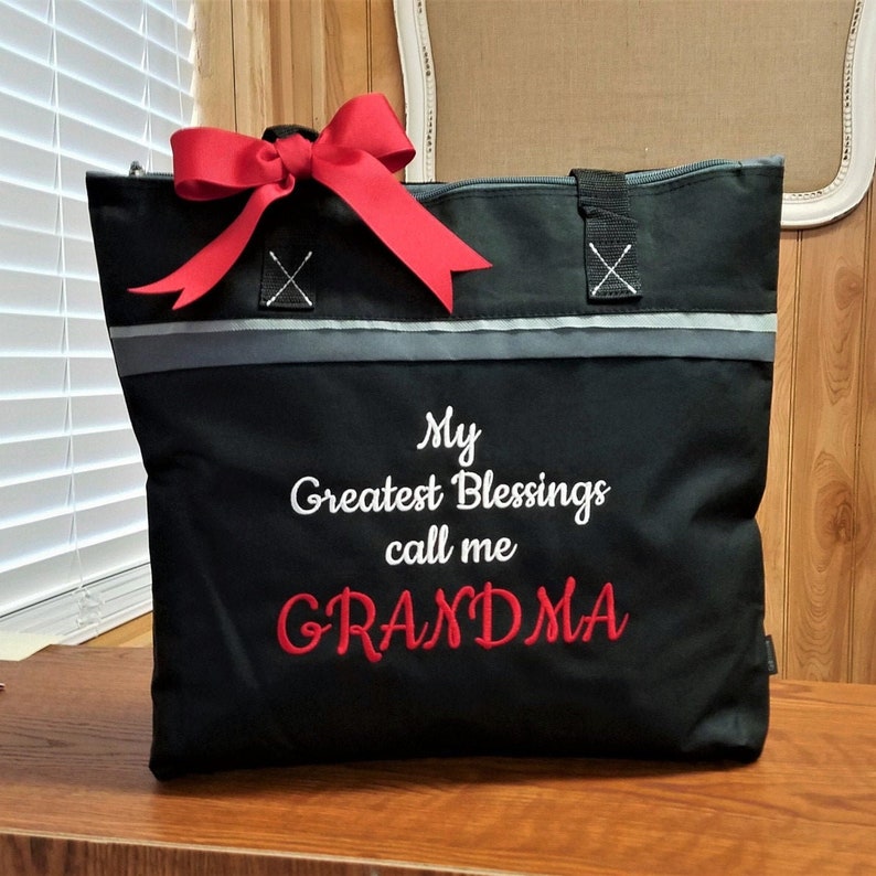 May include: Black zippered tote bag with a red bow. The bag has a gray stripe and the text "My Greatest Blessings call me GRANDMA" embroidered in white.