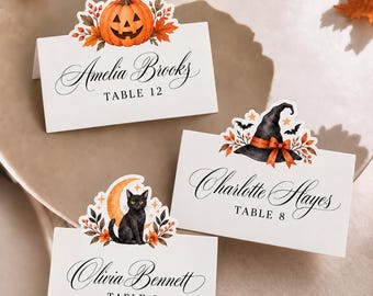 May include: Three white folded place cards featuring Halloween-themed illustrations. One card shows a jack-o'-lantern, one a witch hat with bats, and one a black cat with a crescent moon. Each card displays a name and table number in black text.