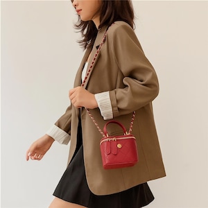 May include: A red, quilted, box-shaped handbag with a gold zipper and handle. The bag has a braided strap and a small gold emblem on the front. The bag is being carried by a person wearing a brown blazer and a black skirt.