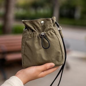 May include: An olive green drawstring bag with a black strap and drawstring closure. The bag has a silver-colored snap closure and metal accents. The bag is held in a person's hand, with a blurred background of trees and a park bench.