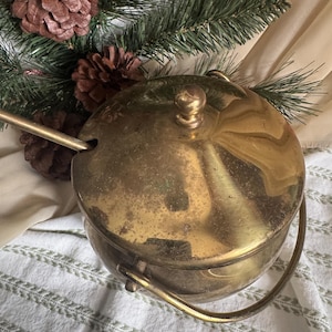 May include: A vintage brass sugar bowl with a lid and handle. The bowl has a round shape and a tarnished finish, suggesting age. A brass spoon rests on the side. The background includes pinecones and evergreen branches, suggesting a holiday theme.