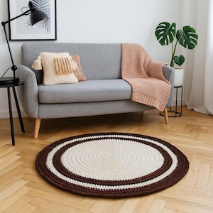 May include: An oval, handmade rug with a cream-colored center and a dark brown border. The rug is made of thick yarn and has a textured appearance. The rug is placed on a wooden floor in front of a gray sofa.