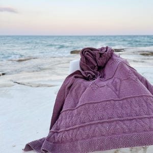 May include: A knitted purple blanket with a textured pattern is draped over a white surface. The blanket features geometric designs and a detailed border. The background shows the ocean and sky, creating a serene and cozy scene.
