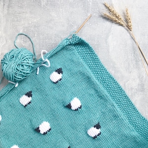 Peut inclure: Une couverture tricotée turquoise ornée de moutons blancs et noirs. Une pelote de laine turquoise et des aiguilles à tricoter sont à côté de la couverture. La couverture est sur une surface grise avec des épis de blé.