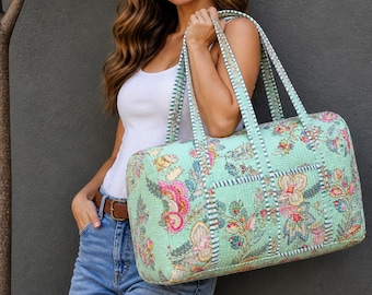 May include: A quilted, mint green duffel bag with a floral pattern in pink, yellow, and blue. The bag has striped handles and trim. The bag is rectangular and appears to be made of a soft fabric.