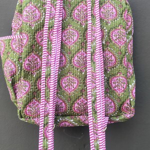May include: A quilted backpack with a green background and a pink floral pattern. The straps and handle are pink and white striped. The backpack has a side pocket and adjustable straps.