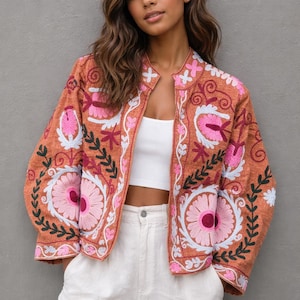 Puede incluir: Chaqueta color óxido con intrincados bordados florales en rosa y blanco. La chaqueta tiene cuello mandarín y se usa sobre una camiseta y pantalones blancos. El bordado incluye grandes patrones de flores y hojas.