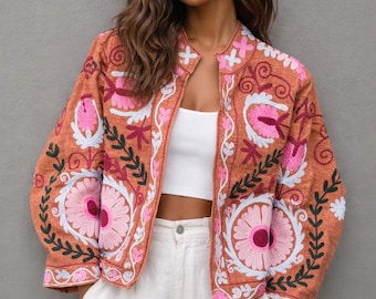 May include: A rust-colored jacket with intricate floral embroidery in pink and white. The jacket features a mandarin collar and is worn over a white top and pants. The embroidery includes large flower and leaf patterns.