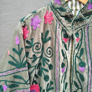 Pode incluir: Uma jaqueta de veludo verde s&aacute;lvia com bordados intrincados em padr&otilde;es florais verde-escuro, rosa brilhante e roxo. A pe&ccedil;a possui gola alta e forro interno vis&iacute;vel com um design de listras brancas e verdes, pendurada em um cabide de madeira.