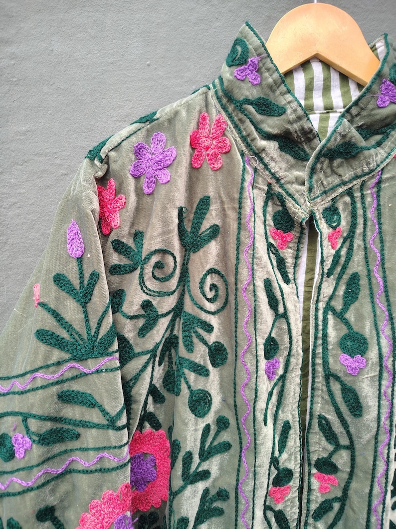 Pode incluir: Uma jaqueta de veludo verde s&aacute;lvia com bordados intrincados em padr&otilde;es florais verde-escuro, rosa brilhante e roxo. A pe&ccedil;a possui gola alta e forro interno vis&iacute;vel com um design de listras brancas e verdes, pendurada em um cabide de madeira.