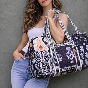 Könnte beinhalten: Eine schwarz-weiß gemusterte Reisetasche mit floralen und geometrischen Designs. Die Tasche hat gestreifte Griffe und eine gesteppte Textur. Die Tasche wird von einer Person getragen, die ein weißes Tanktop und blaue Jeans trägt.