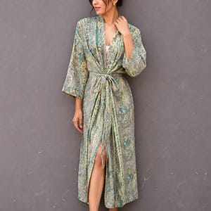 Könnte beinhalten: Ein langer Morgenmantel mit einem detaillierten Paisley-Muster in Salbeigrün, Gelb und Petrol. Das Kleidungsstück hat weite Dreiviertelärmel, einen Schalkragen und einen passenden Bindegürtel aus Stoff, der in der Taille gebunden wird.