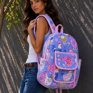 Puede incluir: Una mochila azul claro con un estampado repetido de gallinas coloridas y diseños florales. La mochila tiene dos bolsillos delanteros y ribetes rosas. Las correas y el asa también son rosas y blancas a rayas.