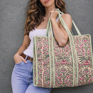May include: A large tote bag with a floral pattern in pink and green on a beige background. The bag has green striped trim and handles. The bag is held by a person wearing a white tank top and blue jeans.