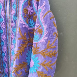 May include: A close-up of a purple jacket with intricate embroidery. The jacket features floral and paisley designs in shades of teal, lavender, and brown. The collar and inner lining have a striped pattern. The jacket is made of a soft, velvety material.