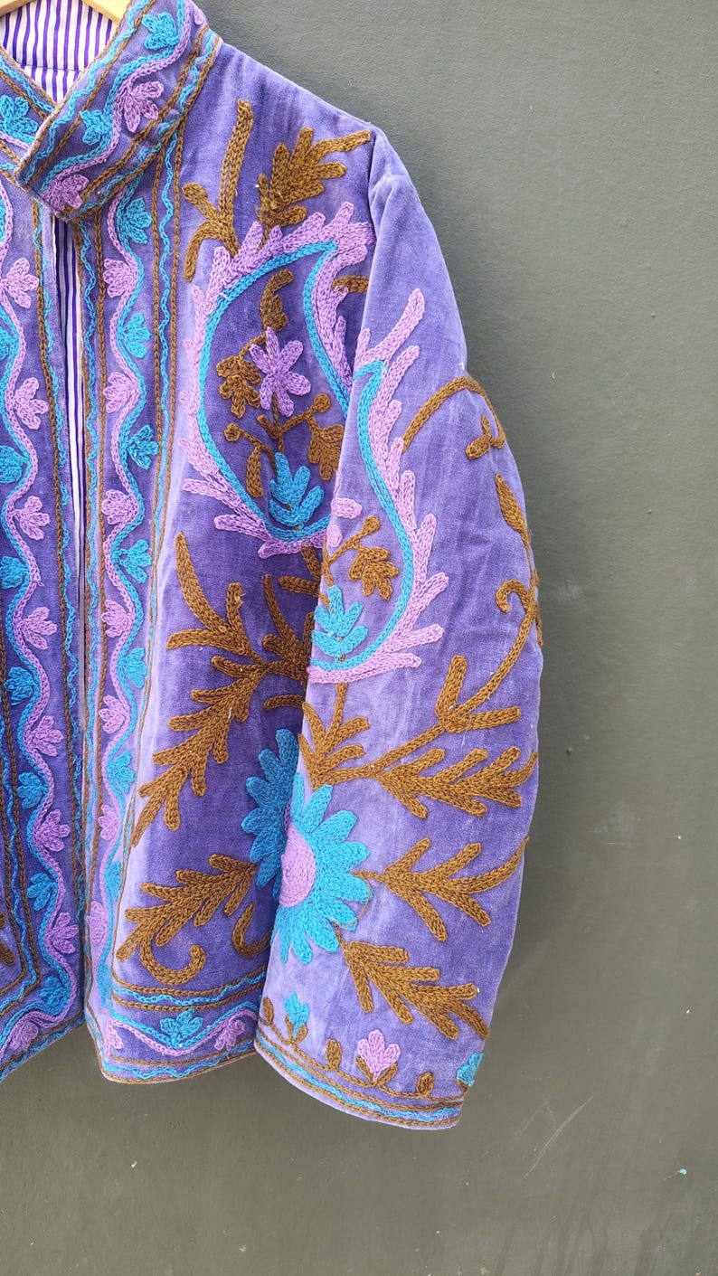 May include: A close-up of a purple jacket with intricate embroidery. The jacket features floral and paisley designs in shades of teal, lavender, and brown. The collar and inner lining have a striped pattern. The jacket is made of a soft, velvety material.