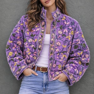 Könnte beinhalten: Gesteppte lila Jacke mit floralen Stickereien in Rosa, Gelb und Grün. Die Jacke hat einen hohen Kragen und einen braunen Gürtel. Das Model trägt eine blaue Jeans und ein weißes Top.