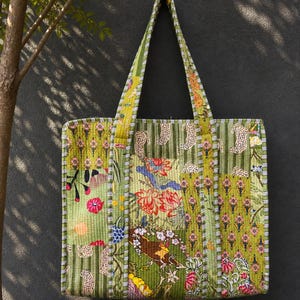 May include: A quilted tote bag with a vibrant floral and animal print in shades of green, red, and yellow. The bag features a striped pattern on the sides and handles, and is hanging against a dark gray wall.