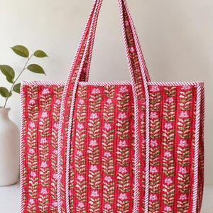 Könnte beinhalten: Eine rechteckige, gesteppte Tragetasche mit leuchtend rotem Hintergrund und einem sich wiederholenden vertikalen Muster aus rosa Blumen und grünen Blättern. Die Tasche hat zwei lange Henkel und Kanten, die mit weiß-rot gestreiftem Stoff eingefasst sind.