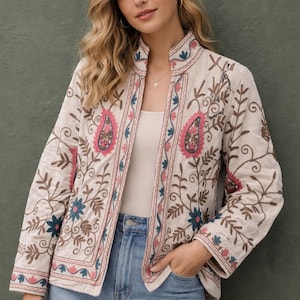 May include: Cream embroidered jacket with a stand-up collar. The jacket has detailed floral and paisley patterns in pink, blue, and brown. It is worn over a white top and blue jeans.