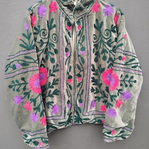 Pode incluir: Uma jaqueta de veludo verde s&aacute;lvia com mangas compridas, gola alta e fecho frontal com z&iacute;per. A pe&ccedil;a &eacute; decorada com bordados intrincados em verde escuro, rosa brilhante e roxo, exibindo padr&otilde;es florais e de vinhas por toda a parte.