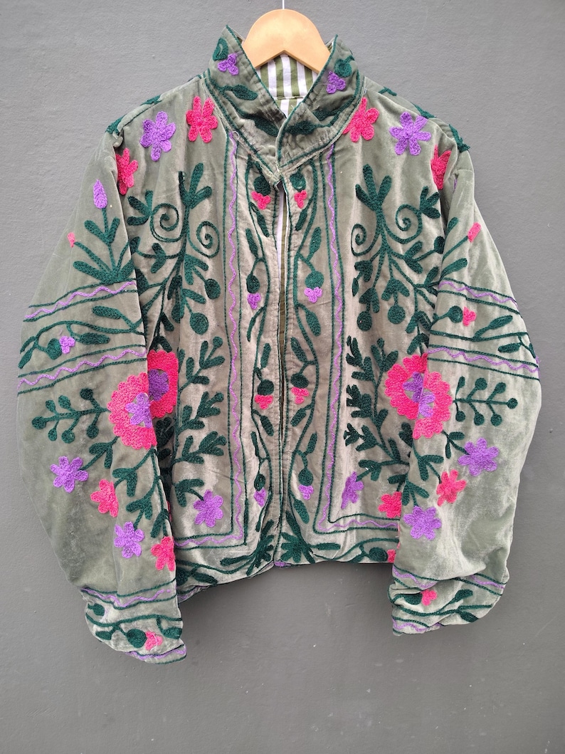 Pode incluir: Uma jaqueta de veludo verde s&aacute;lvia com mangas compridas, gola alta e fecho frontal com z&iacute;per. A pe&ccedil;a &eacute; decorada com bordados intrincados em verde escuro, rosa brilhante e roxo, exibindo padr&otilde;es florais e de vinhas por toda a parte.