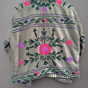 Pode incluir: Uma jaqueta verde-s&aacute;lvia com bordados florais em verde escuro, rosa choque e roxo. O design inclui um grande motivo floral central, faixas horizontais com vinhas e pequenas flores. A jaqueta tem gola alta e forro listrado.