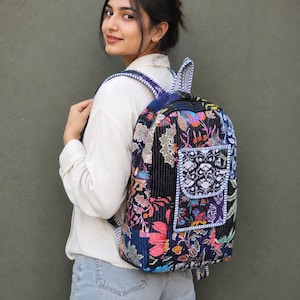 Könnte beinhalten: Ein gesteppter Rucksack mit dunkler Basis und einem lebendigen, mehrfarbigen Blumen- und Pflanzenmuster. Die Tasche verfügt über eine Vordertasche mit Klappenverschluss, verstellbare Schultergurte und sichtbare Nähte auf der Stoffoberfläche.