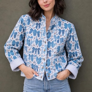 Könnte beinhalten: Blau-weiß gemusterte Jacke mit Stehkragen und Knopfverschluss. Die Jacke hat ein sich wiederholendes Blumenmuster und wird mit hellblauen Jeans kombiniert. Die Manschetten haben ein Streifenmuster.