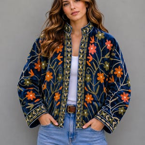 Könnte beinhalten: Marineblaue Samtjacke mit orangefarbener und grüner Blumenstickerei. Die Jacke hat einen Stehkragen und ist vorne offen, wodurch ein weißes Oberteil und ein brauner Gürtel sichtbar werden. Die Ärmel sind lang, und die Jacke wird mit einer Jeans getragen.