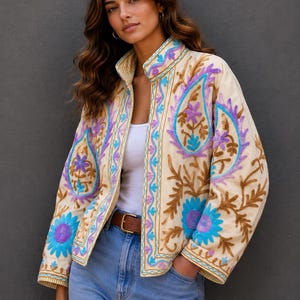 May include: Cream-colored embroidered jacket with a high collar. The jacket features intricate floral and paisley designs in shades of blue, purple, and brown. A brown belt cinches the waist, and the model is wearing blue jeans.